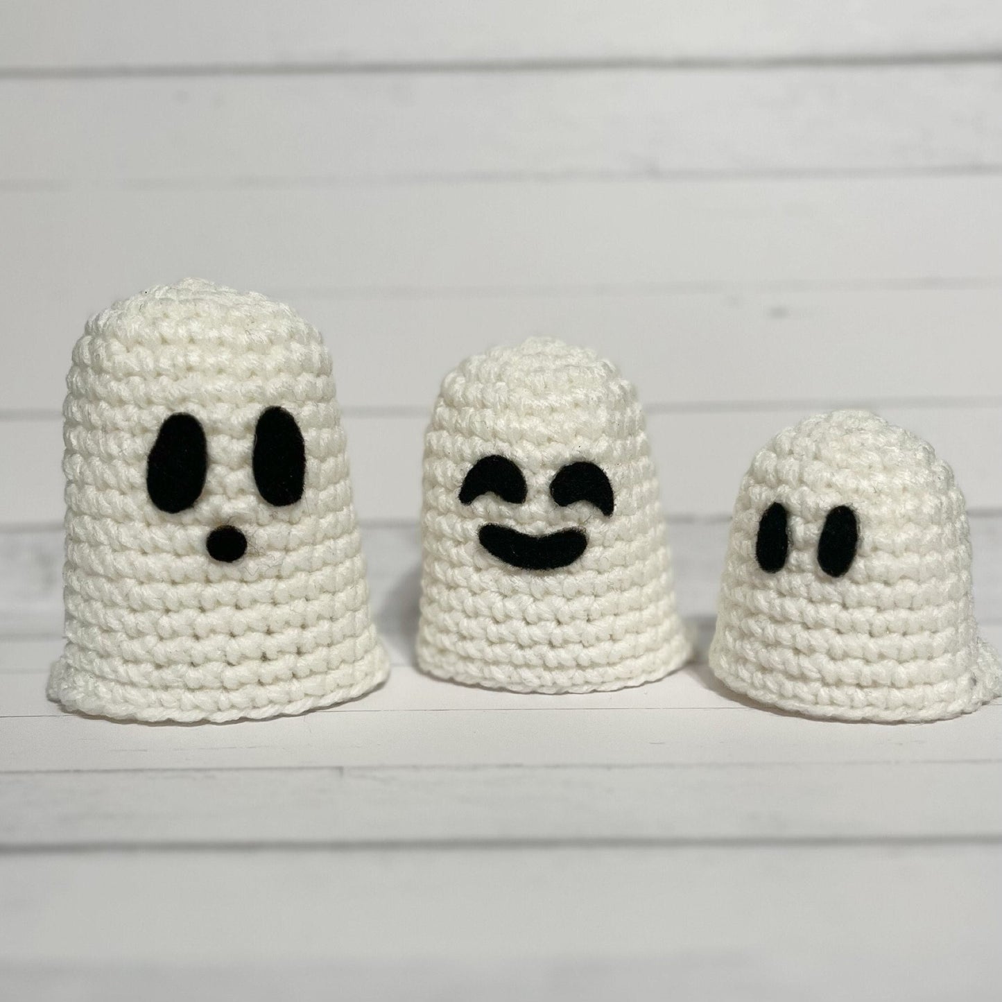 three white crocheted ghosts. The largest is 4" high and has 2 black oval eyes and a round nose. The middle size has 2 black laughing eyes and black smiling mouth and stands 3.5". The smallest is 2.5" high and has 2 black oval eyes